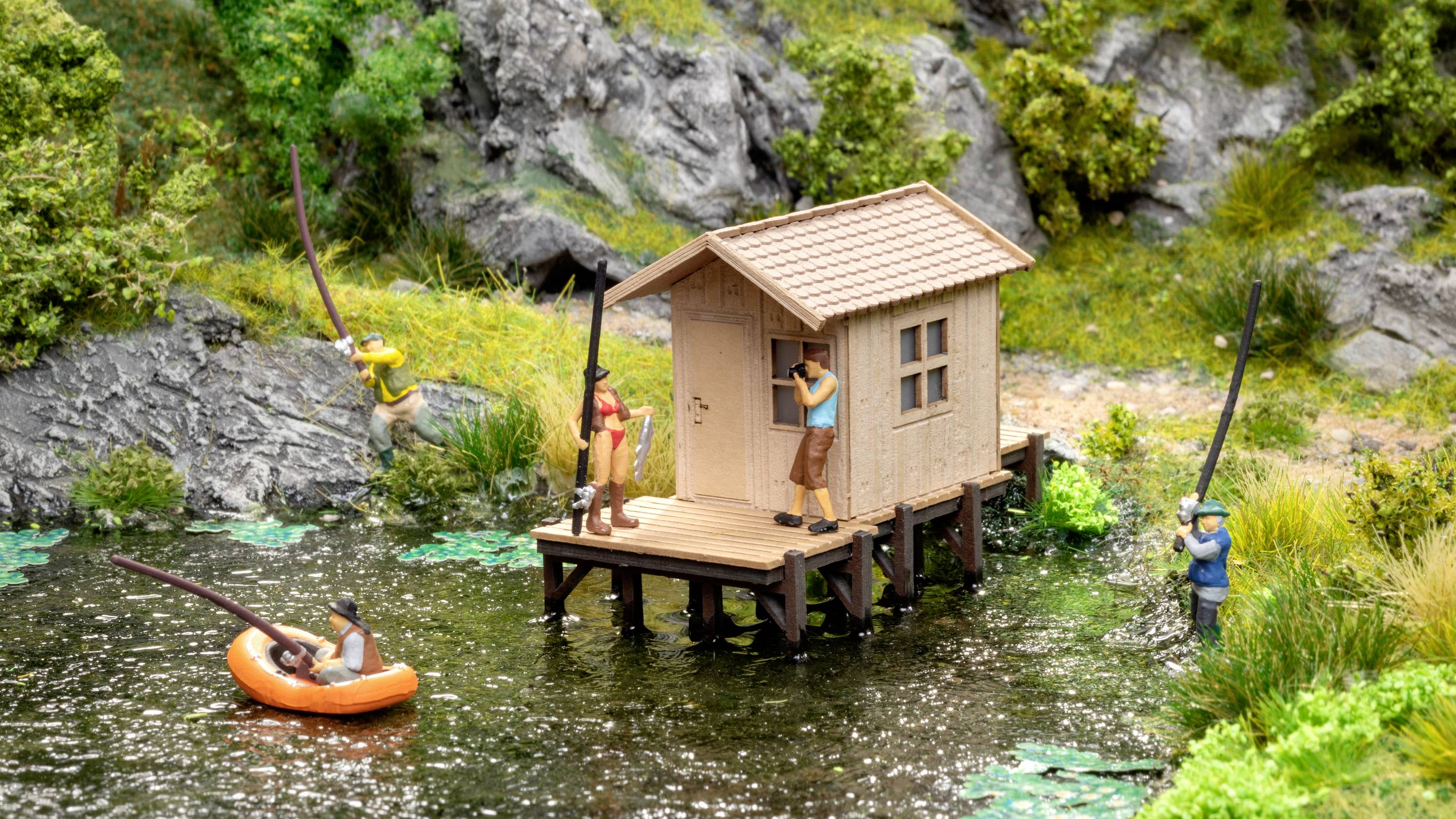 A miniature model shows a wooden house on stilts by the lakeshore, surrounded by rocks and greenery. Figures are fishing from the jetty and from an inflatable boat.