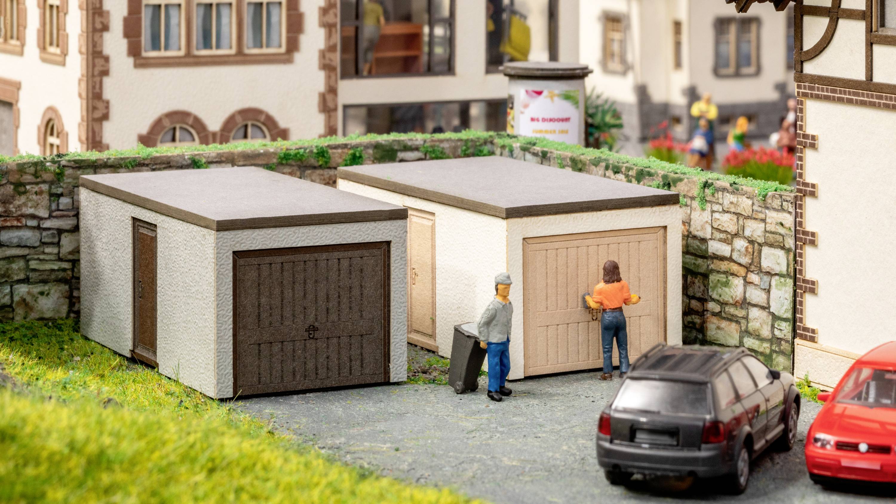 Two garages in a miniature town. One person stands in front of the right-hand garage. A black car is parked in front. Surrounded by model houses.
