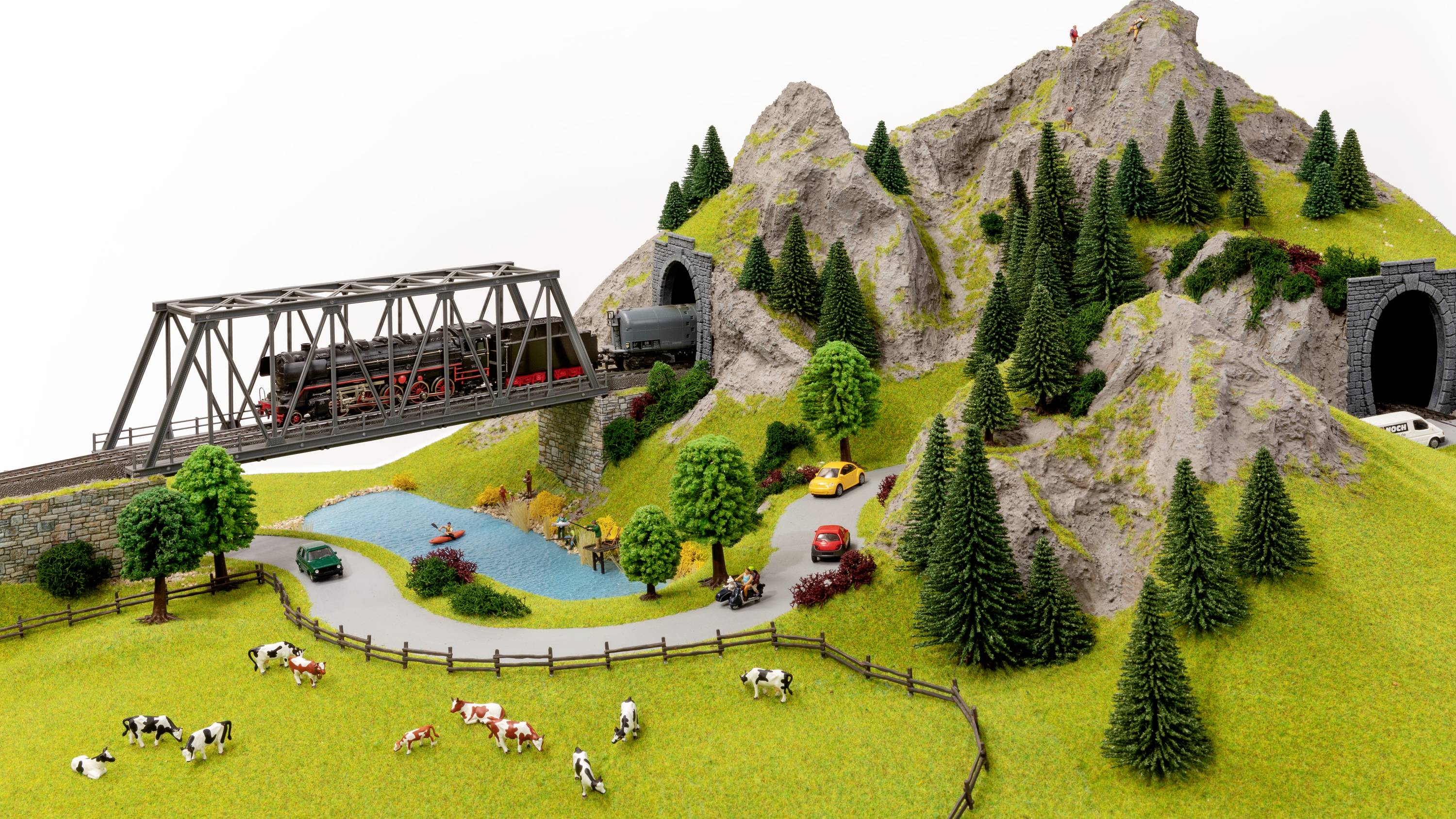 A model railway travels over a bridge through a mountainous landscape with tunnels. Cows are grazing on a meadow beside a lake.