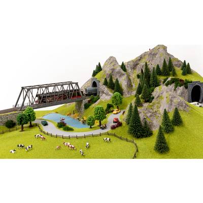 A model railway travels over a bridge through a mountainous landscape with tunnels. Cows are grazing on a meadow beside a lake.