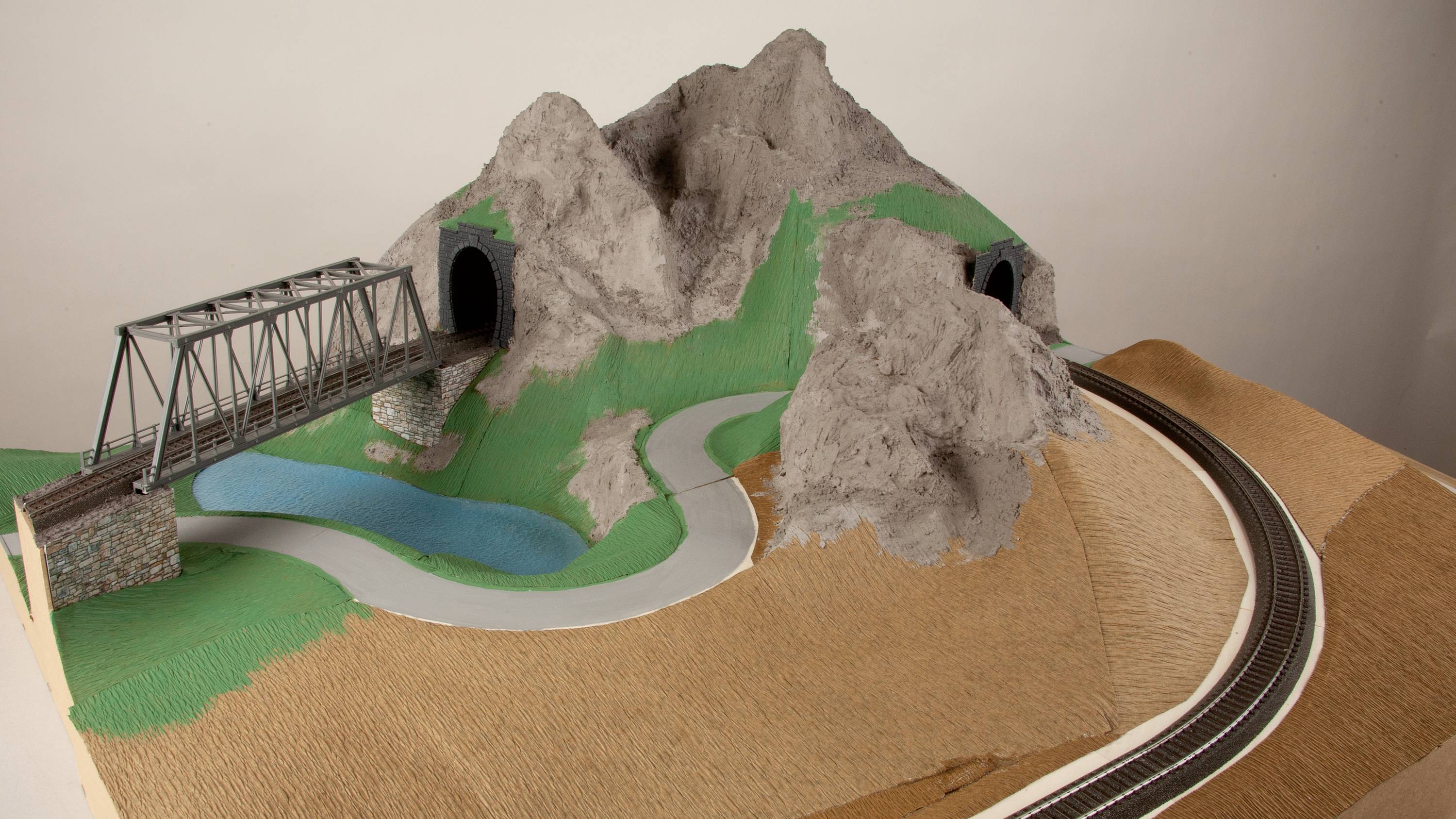 A model of a railway landscape with a bridge and two tunnels in a mountainous terrain, surrounded by a road and a pond.