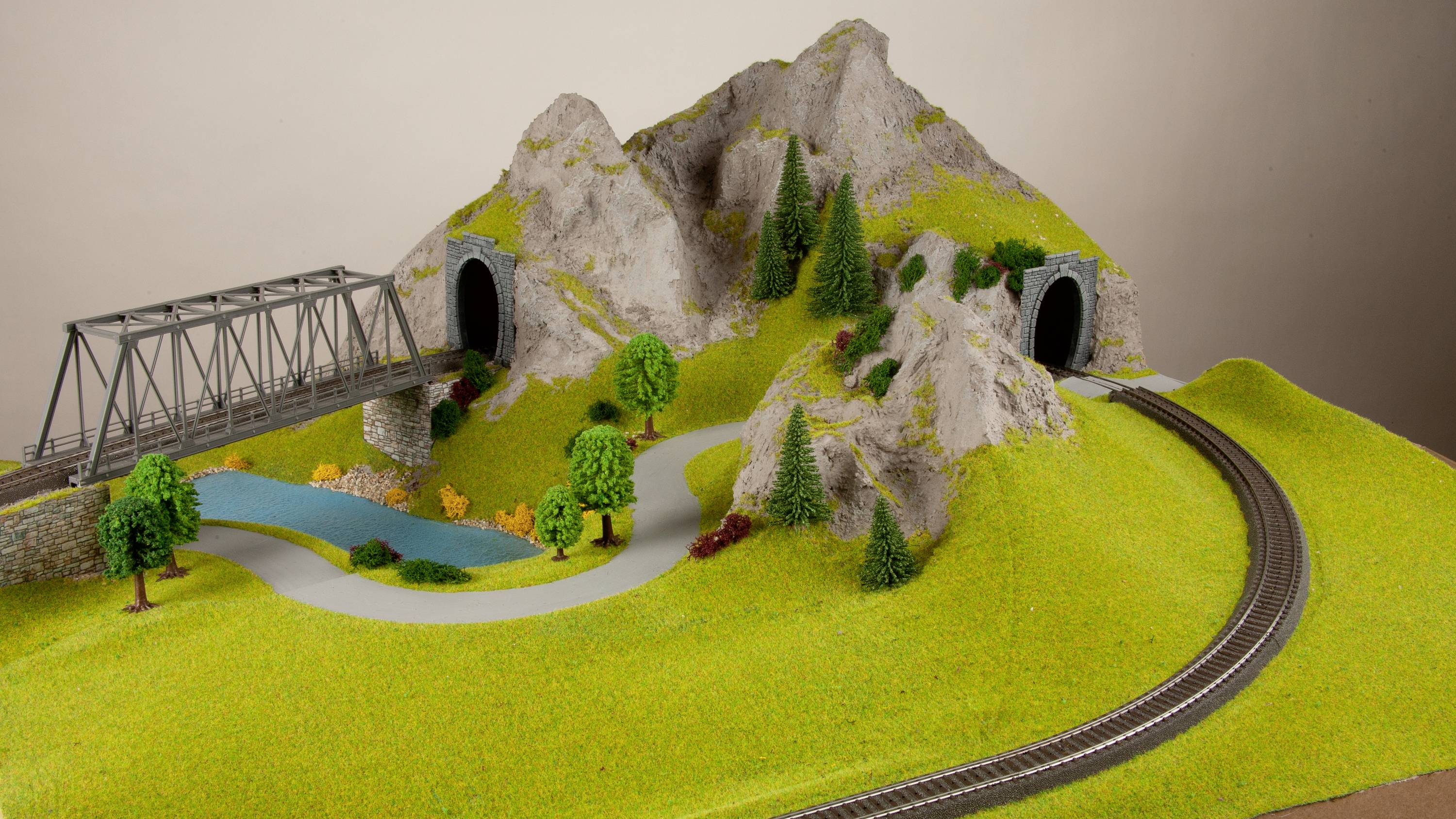 Model railway landscape with a bridge, two tunnels in hills, forest, meadows and railway tracks; shows a realistic miniature scene.