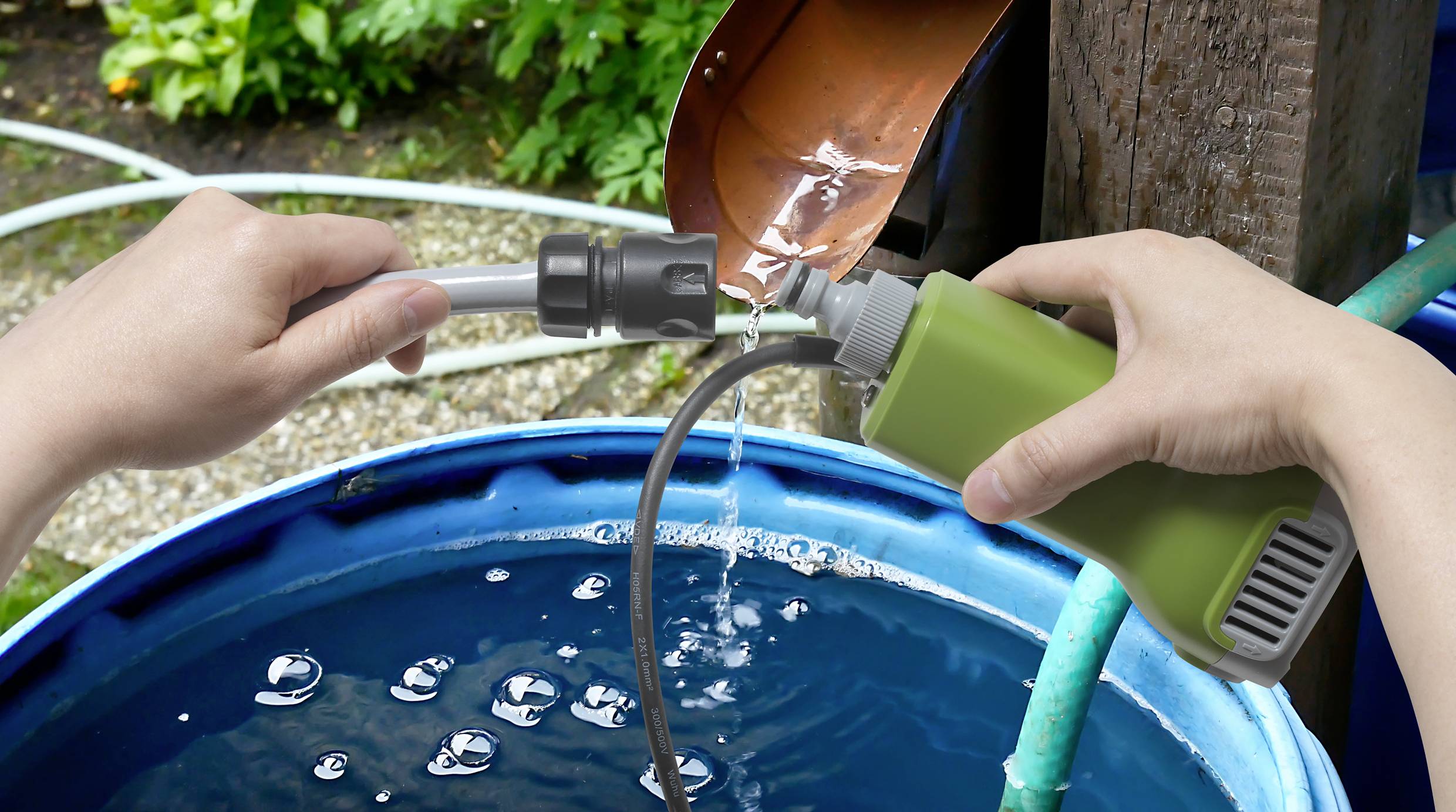 A person is using a flexible hose to fill a blue water butt outdoors, collecting water from a rainwater downpipe.