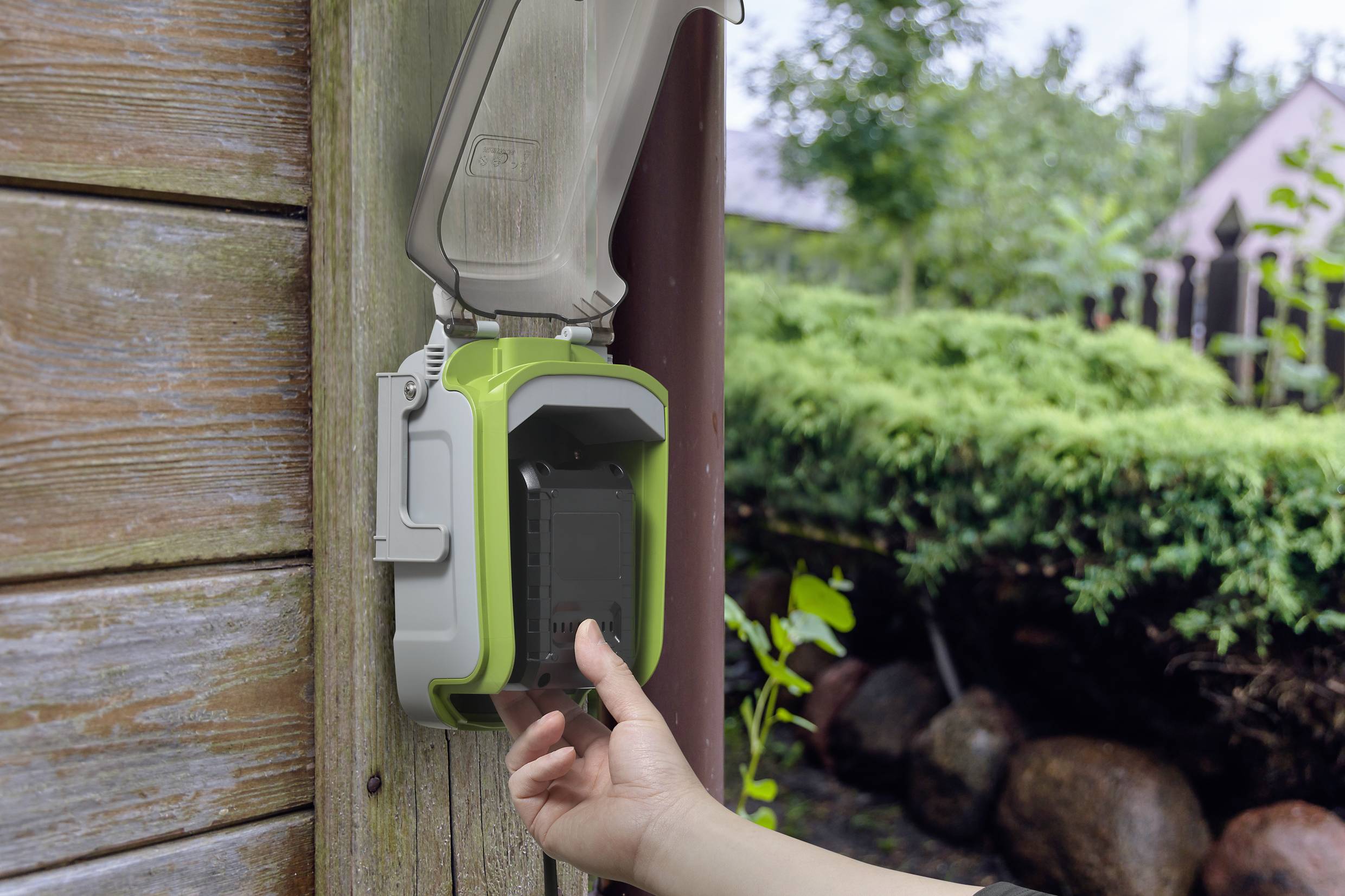 A person plugs a smartphone into an outdoor charger on a wooden house wall. A garden is visible in the background.