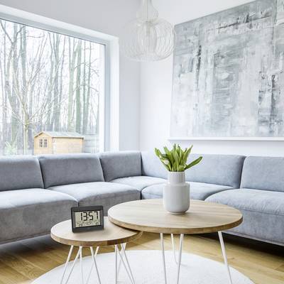 Bright living room with a grey sofa, round wooden table and digital clock. Large wall art and windows overlooking trees.