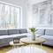Bright living room with a grey sofa, round wooden table and digital clock. Large wall art and windows overlooking trees.