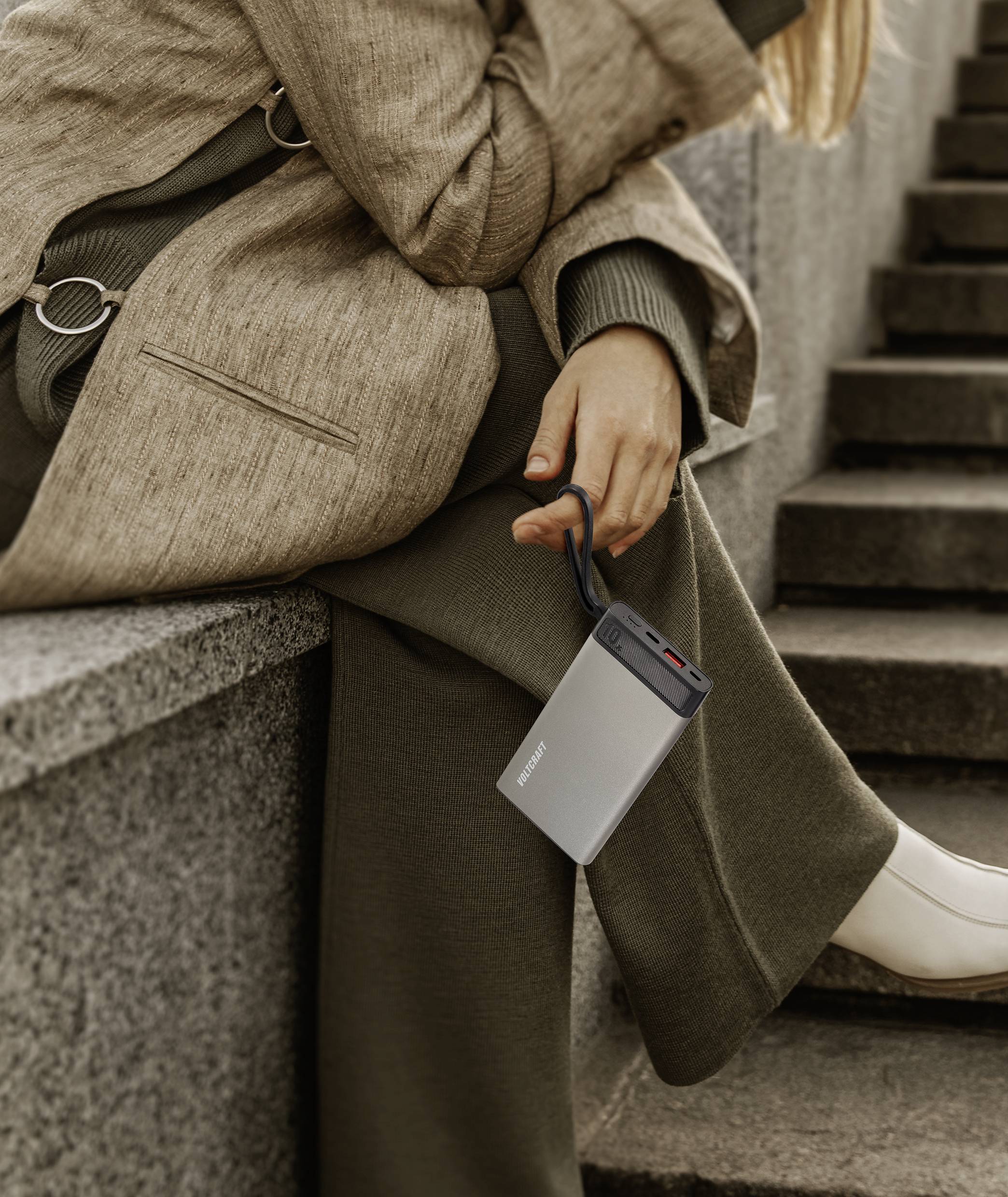 A person is sitting on stone steps, holding a portable, silver charger with a cable.