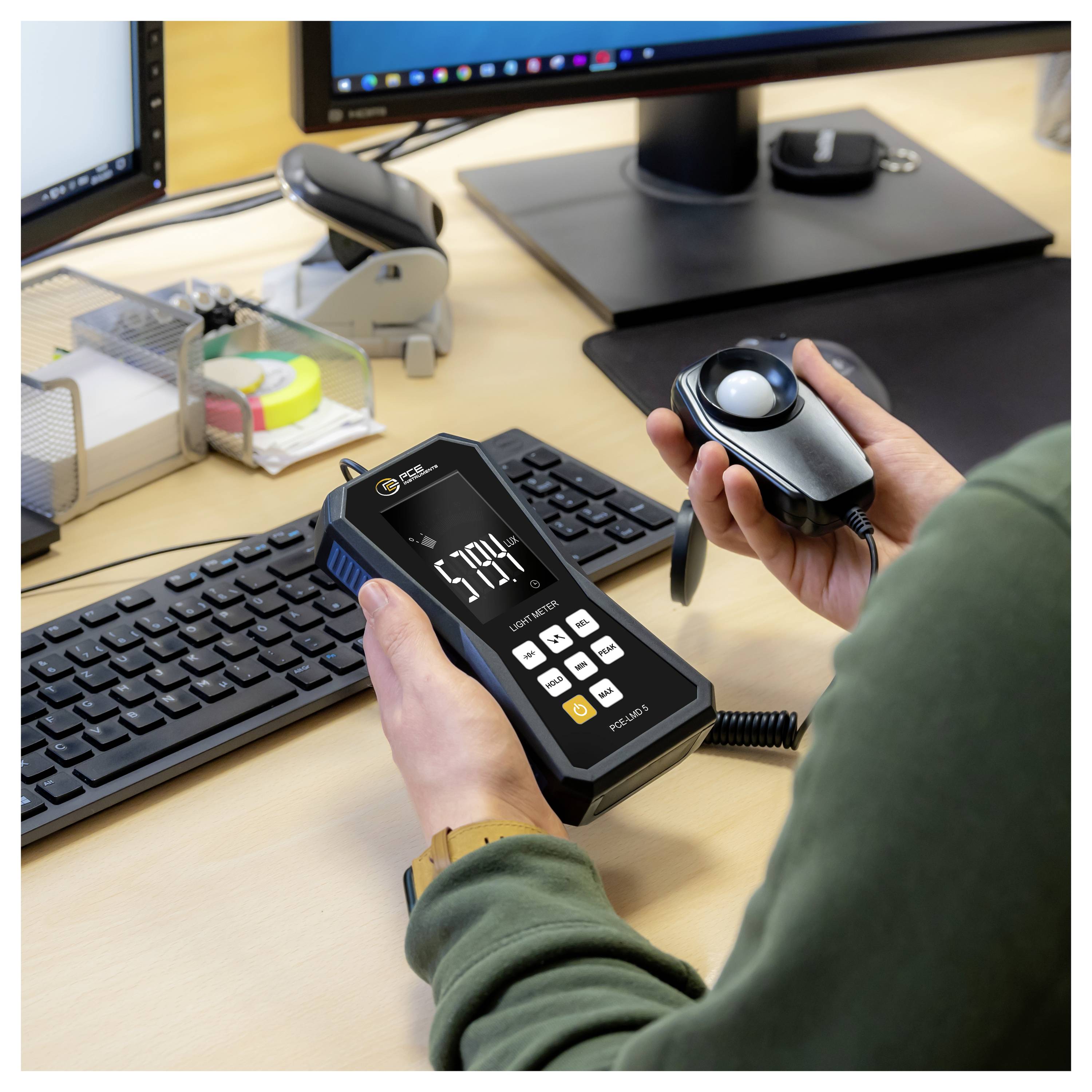 Person using a handheld light meter and wired sensor at a desk, measuring screen brightness near a computer monitor and keyboard.