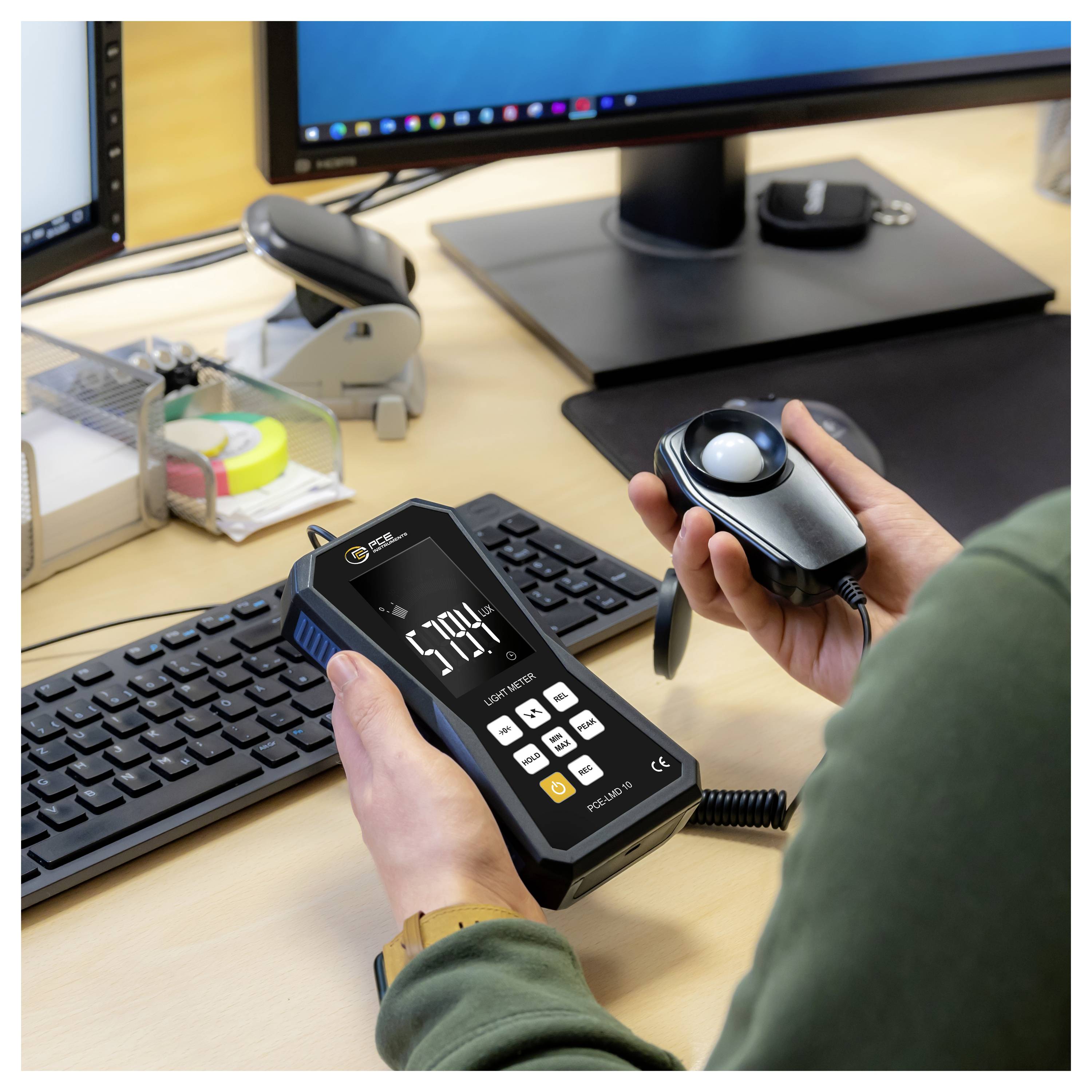 Person holding a digital meter and a handheld light sensor at a desk, measuring monitor brightness in an office setup.