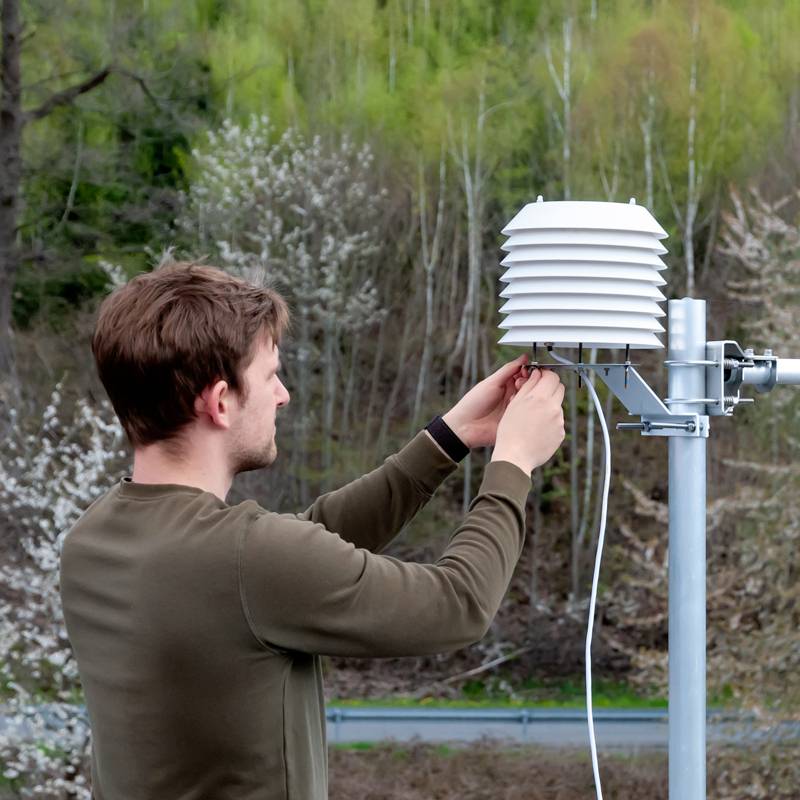 A man is checking a weather station outdoors, surrounded by trees. The weather station has a round, stacked sensor.