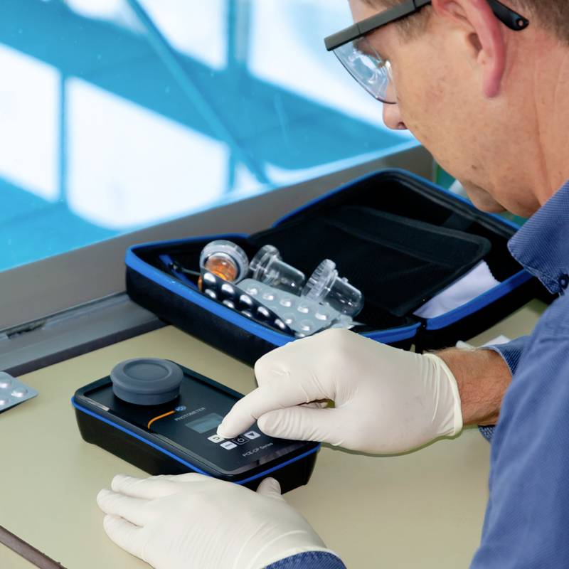 A person wearing laboratory gloves is using a portable photometer on a table. Laboratory equipment and solutions are visible in the background.