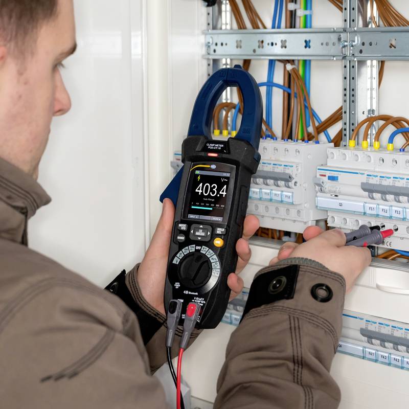An electrician is measuring the voltage in a fuse box with a multimeter. Cables and fuses are visible.