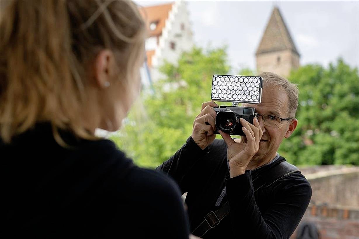 A person is photographing another person outdoors with a camera that has a flash. Trees and buildings are visible in the background.