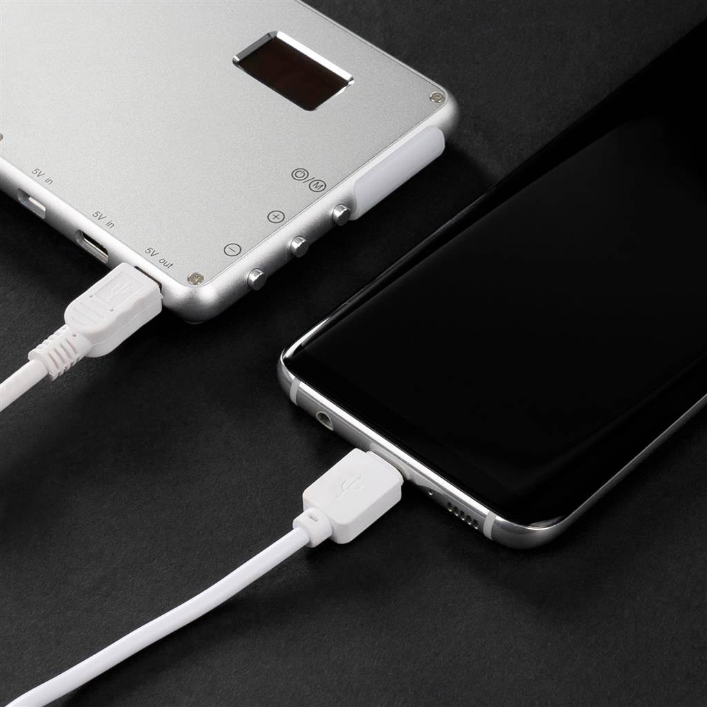 A smartphone is connected to a portable silver power bank with a white cable. Both devices are resting on a dark surface.