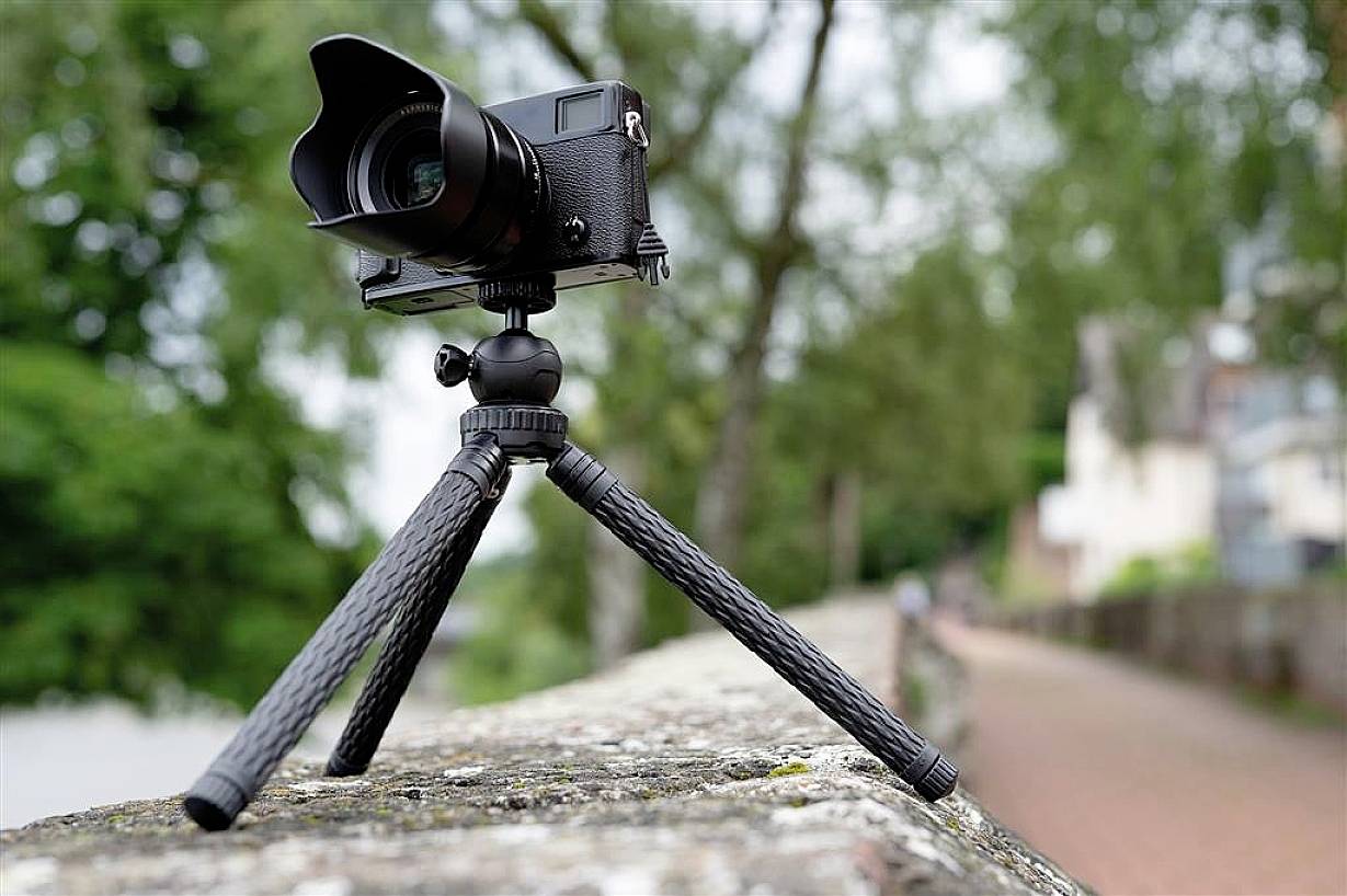 A camera is mounted on a tripod, positioned on a brick wall outdoors. Trees and a path are visible in the background.