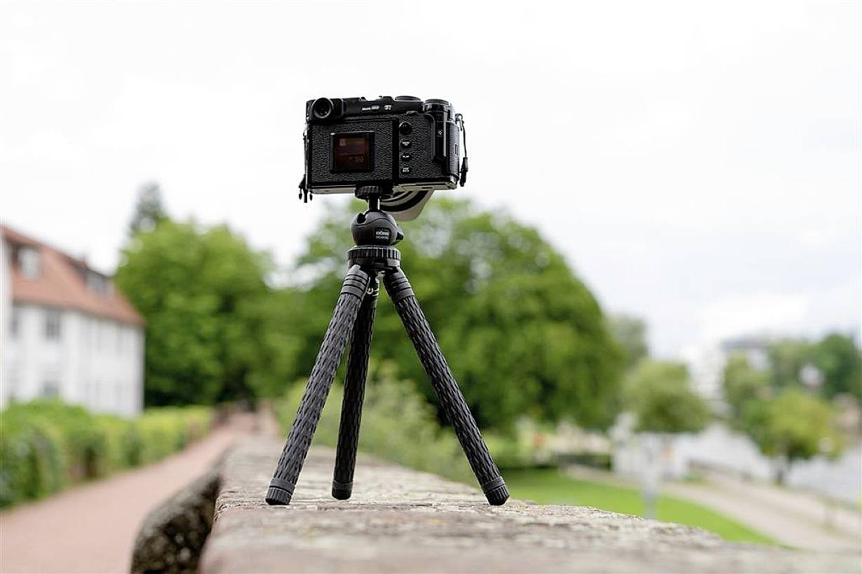 A black camera on a tripod stands on a stone wall with a blurry background of trees and buildings.