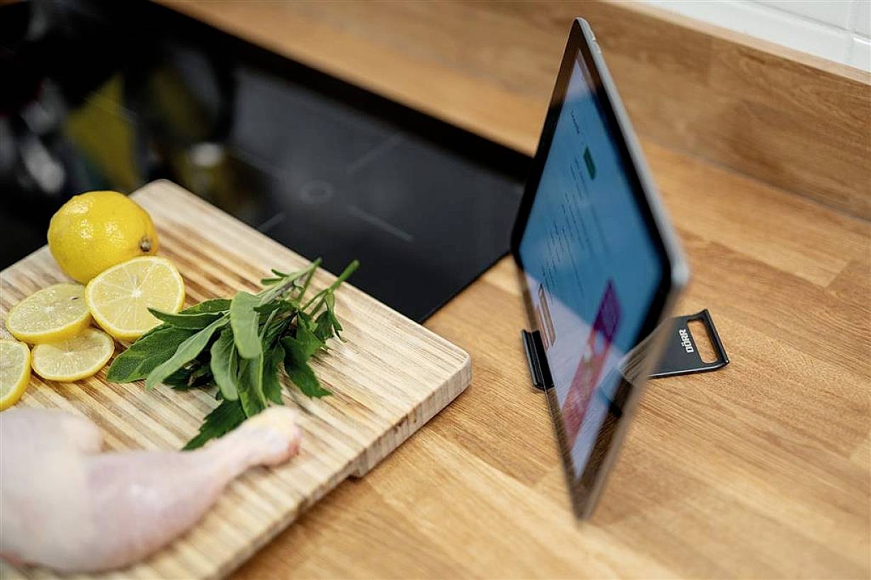 A tablet is standing on a kitchen worktop, displaying a recipe. Beside it lies a chopping board with lemons, herbs, and a chicken leg.