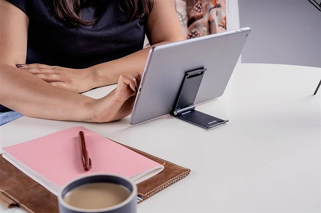 A person is tapping on a tablet, which is mounted on a stand on a table. Beside it lie a notebook and a cup of coffee.