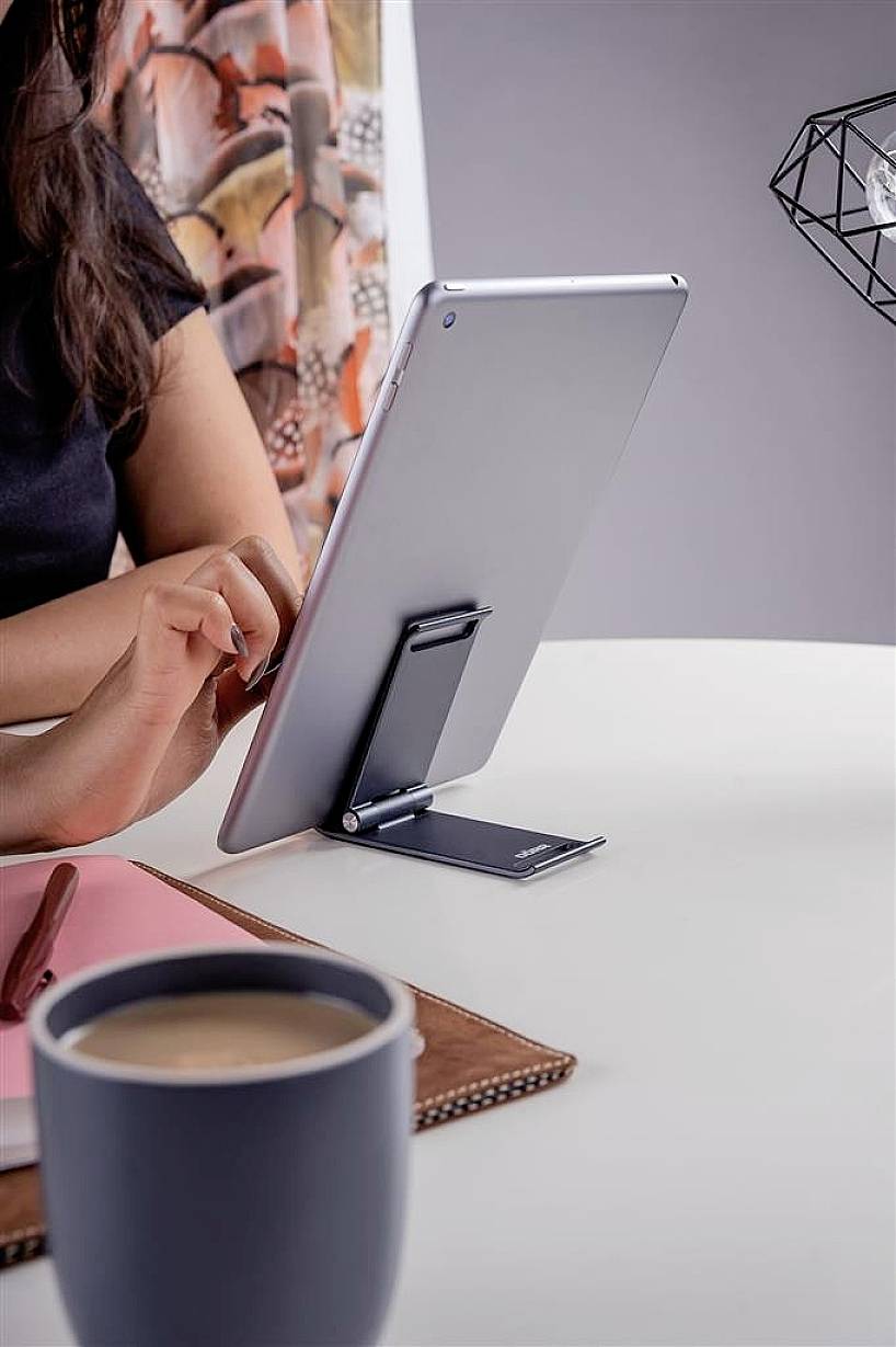 A person is typing on a tablet, which is positioned on a stand on a table. Notebooks and a cup of coffee are placed beside the tablet.