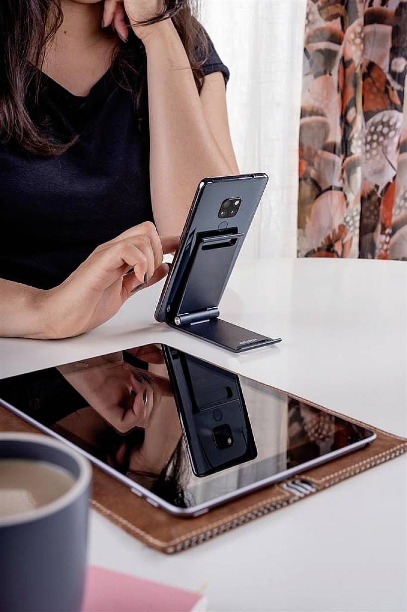 A woman at a table uses a smartphone on a stand, with a tablet beside it. A cup of coffee sits in the foreground.