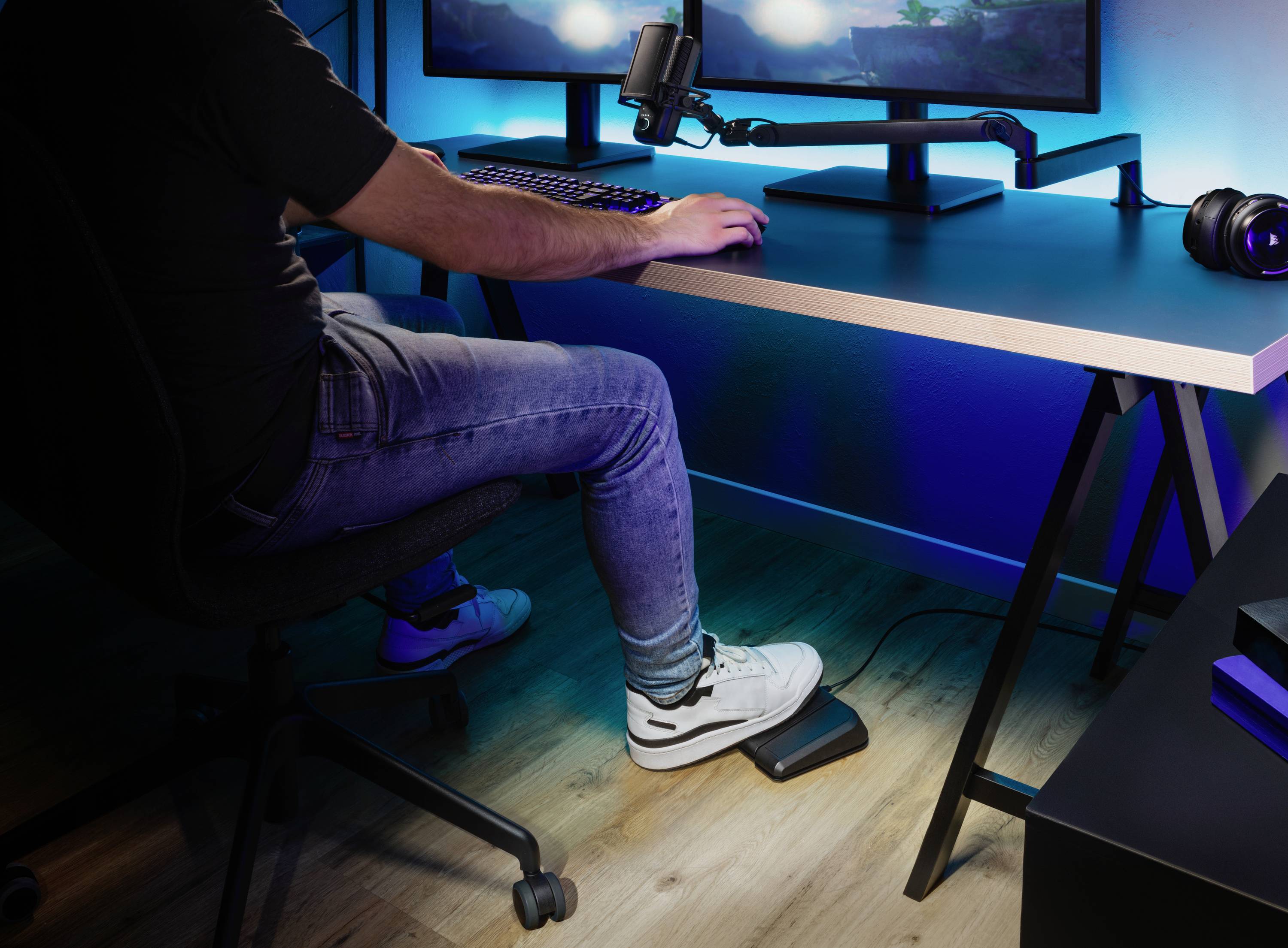 A person is sitting at a desk with a monitor and microphone, wearing headphones and operating a pedal with their foot underneath the desk.