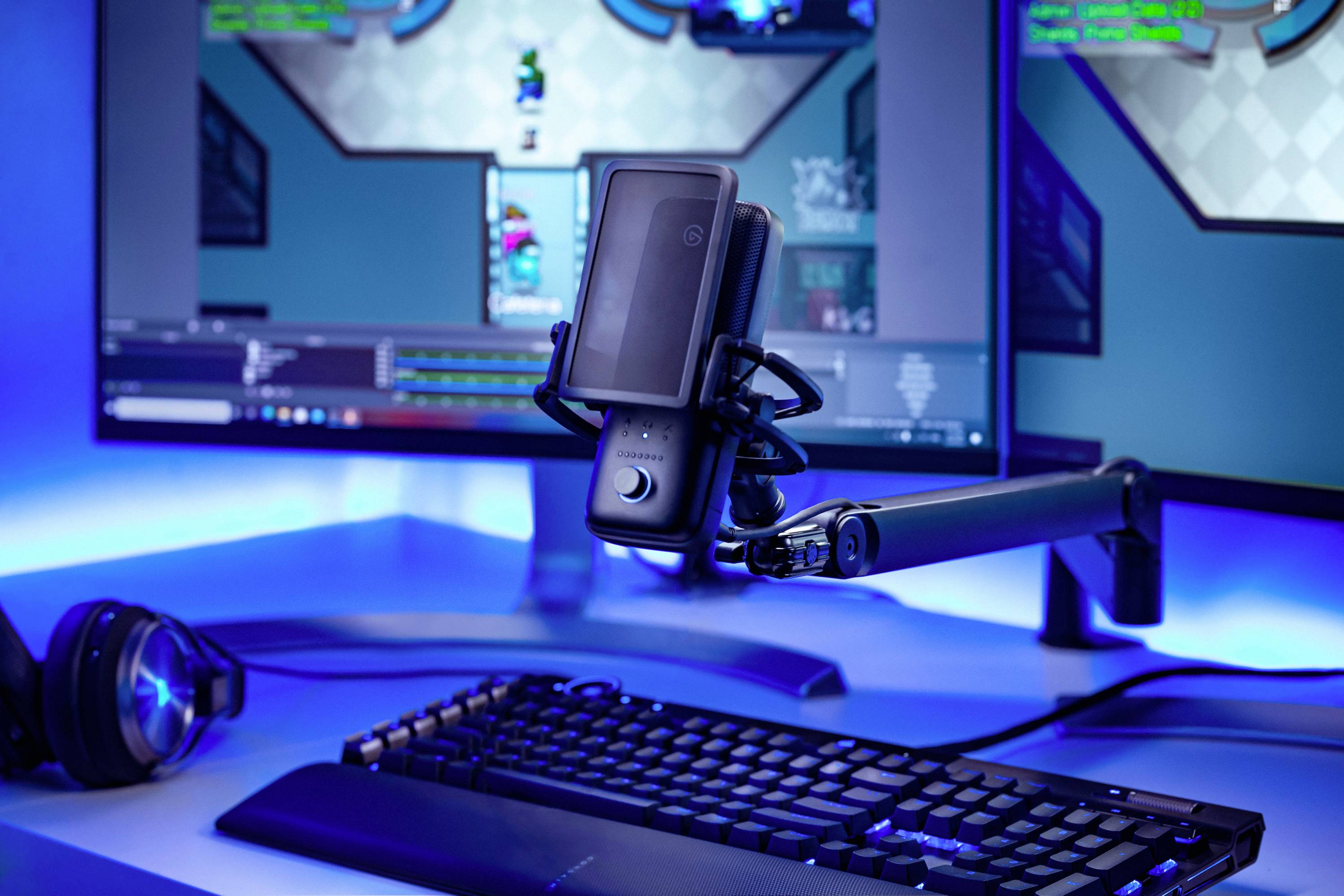 A computer setup with a keyboard, microphone, and headphones. Two screens with a video game are visible in the background.