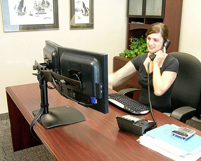 A woman is sitting at a desk, talking on the phone and working on a computer. The desk contains office supplies and folders.