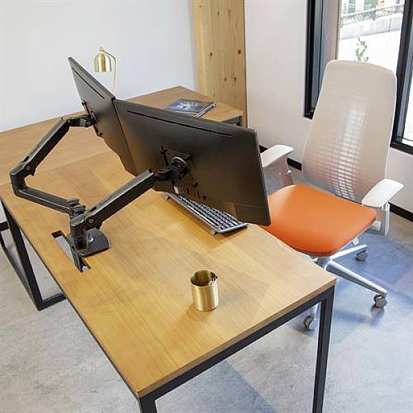 Two monitors on an adjustable stand on a wooden desk, next to an empty office chair, a coffee mug, and a notebook.