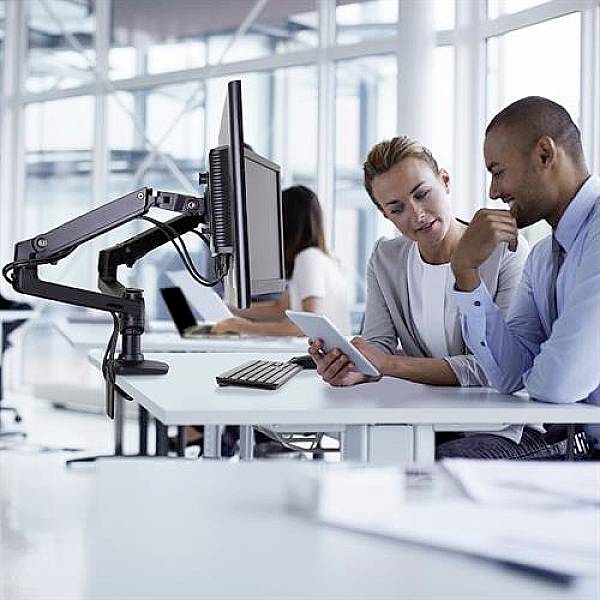 Two people in an office, looking together at a tablet, with computers in the background. They appear focused and are discussing something.
