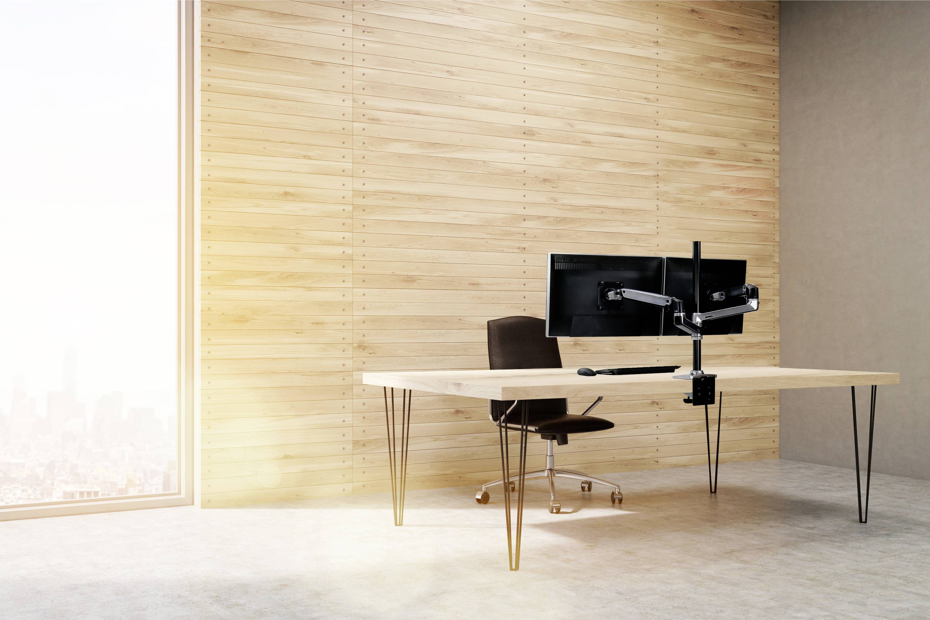 Modern desk with two monitors on a monitor arm and a black chair. The room has wooden walls and a large window view.