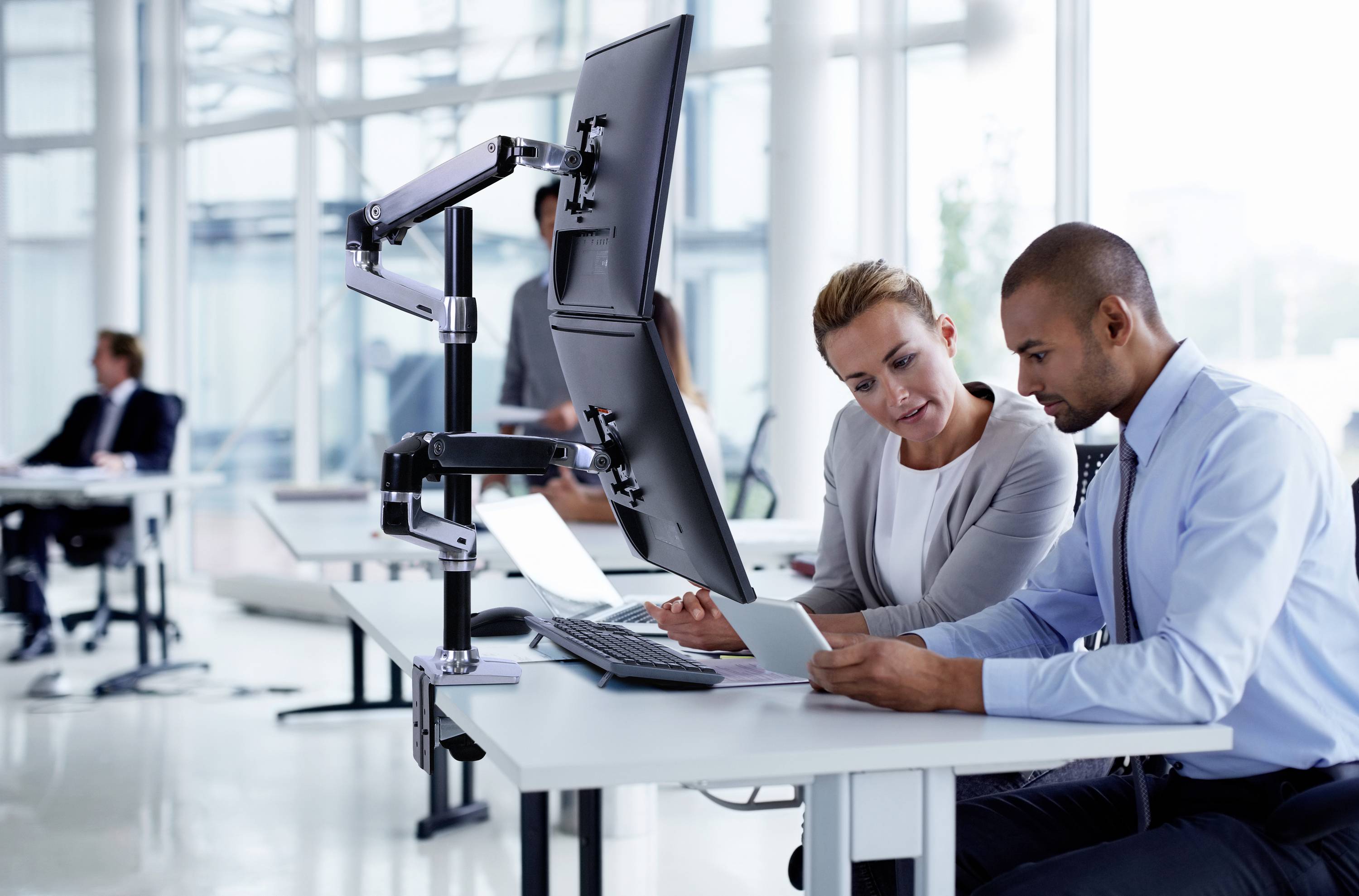 A man and a woman are sitting at a desk in a modern office, looking at a tablet. Behind them are work colleagues.