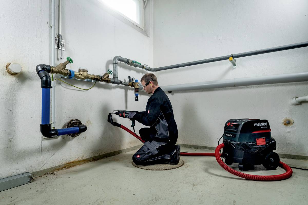 A person in work attire is operating a device in a technical room. A vacuum cleaner is connected to them via a hose.