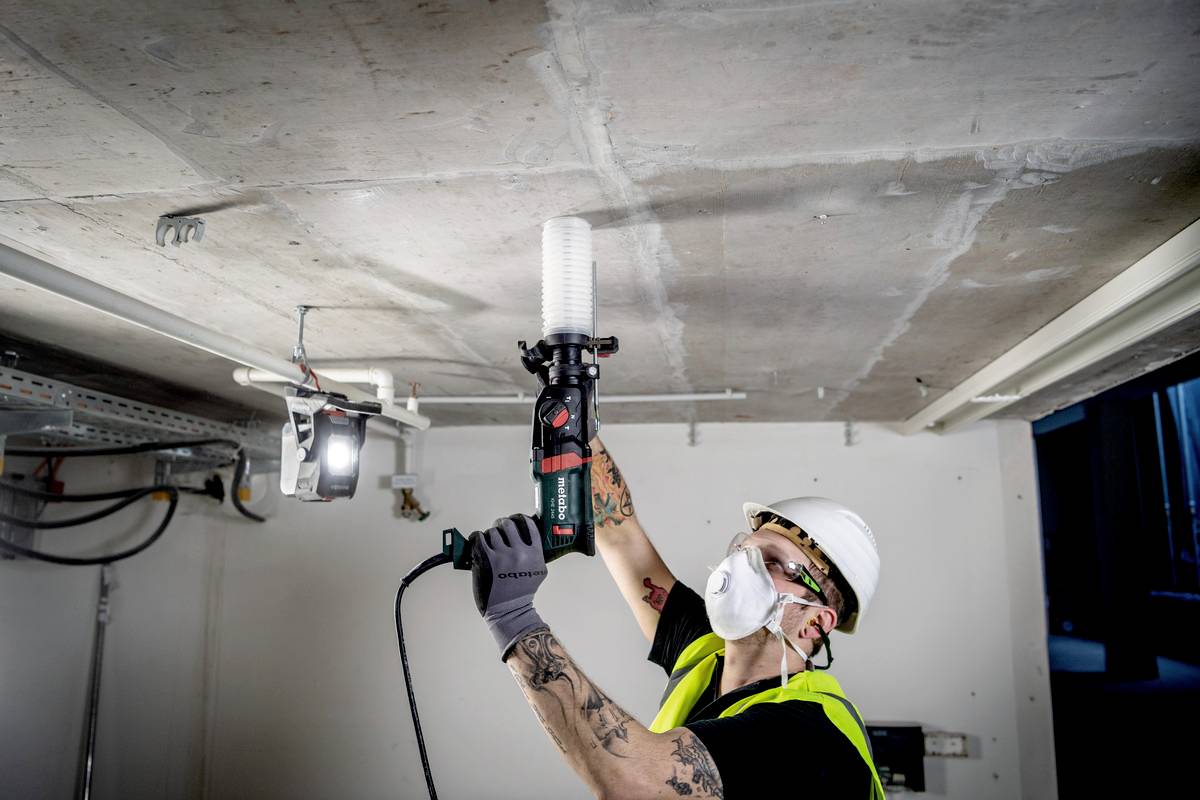 A construction worker in safety clothing and a respirator mask is drilling into a concrete ceiling with an electric drill.