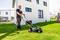A man is mowing the lawn with a lawnmower in front of a modern, white house. Other buildings can be seen in the background.