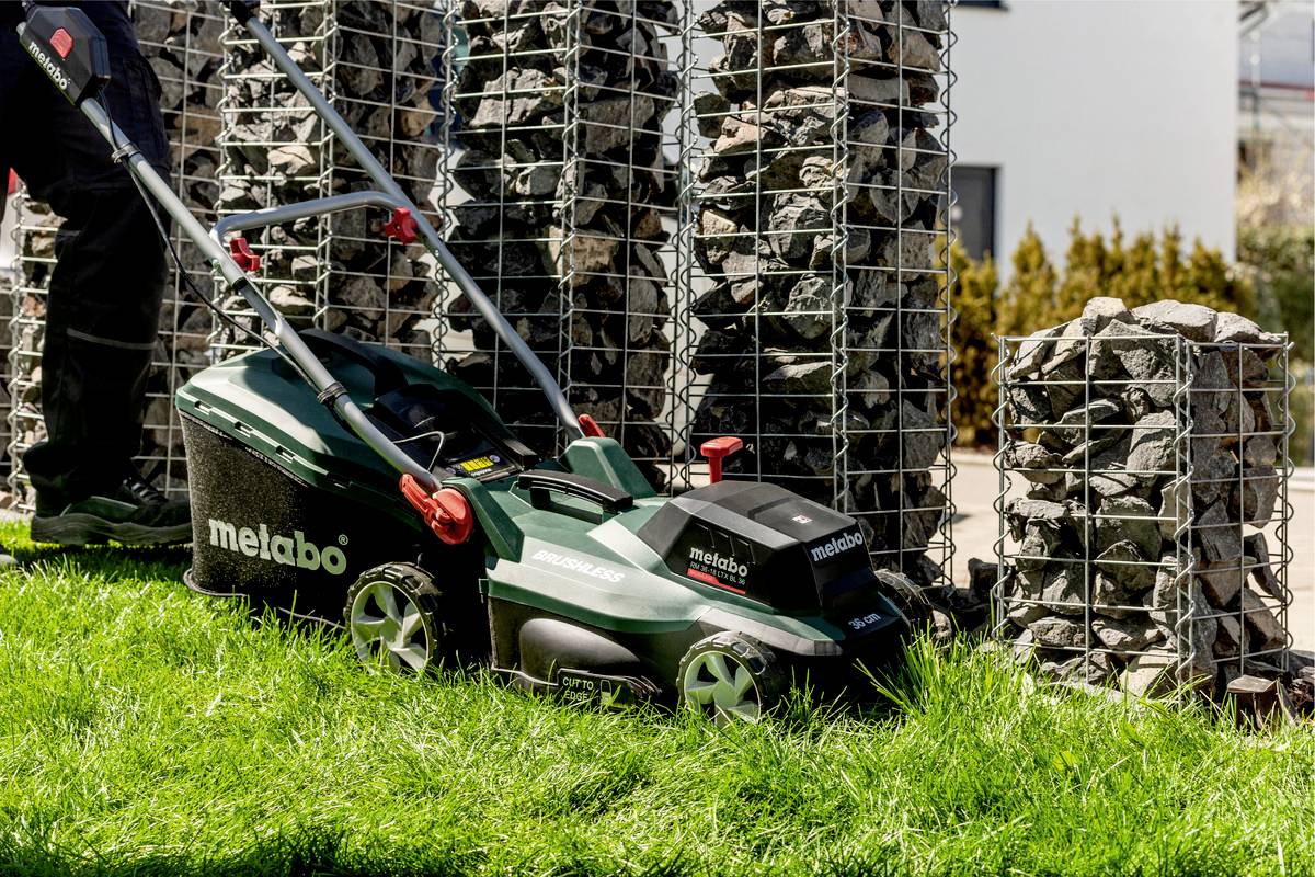 A person is mowing with a green lawnmower on a grassy area next to gabion walls filled with stones.