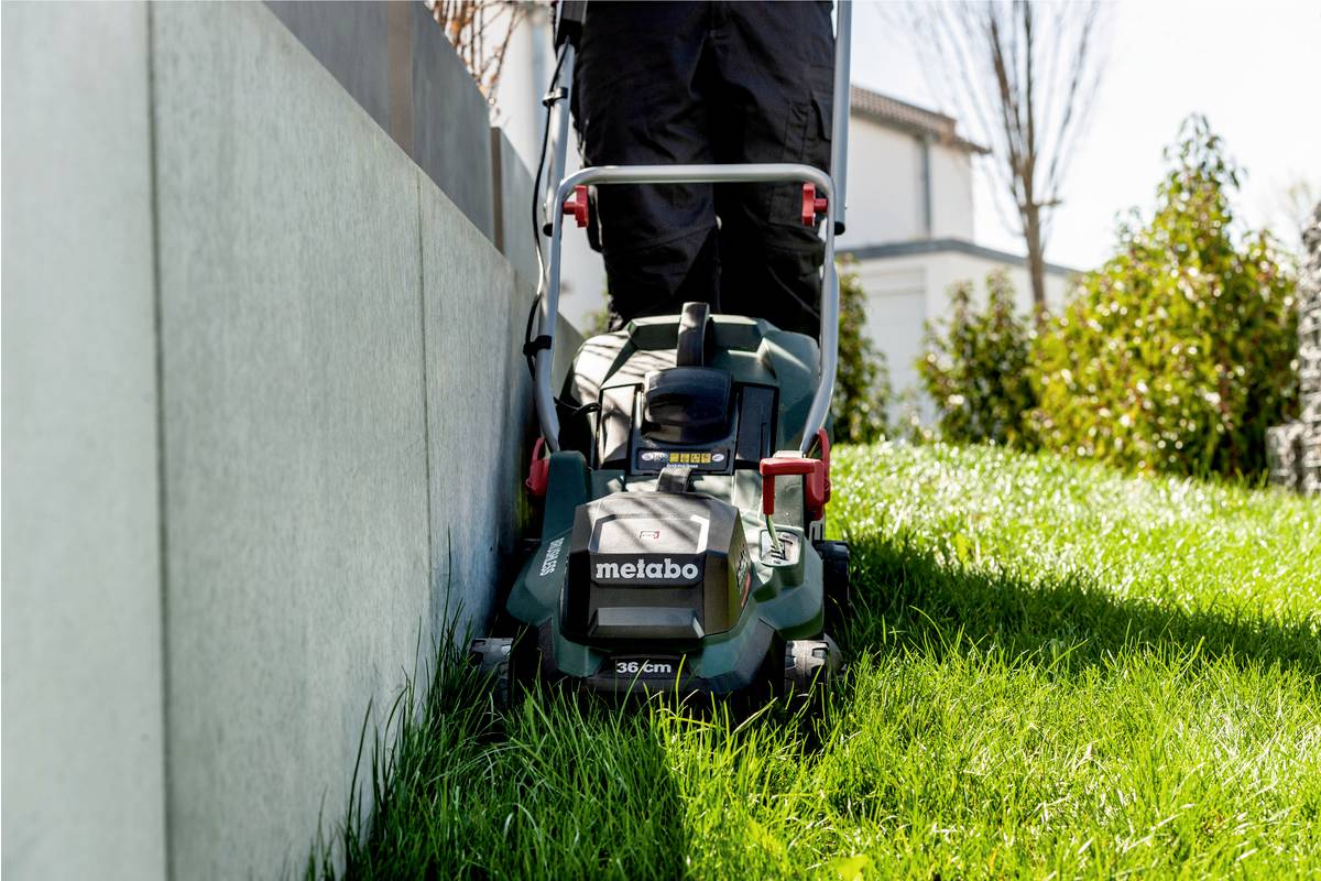 A person is mowing the lawn with a green lawnmower alongside a concrete wall in a sunny garden.
