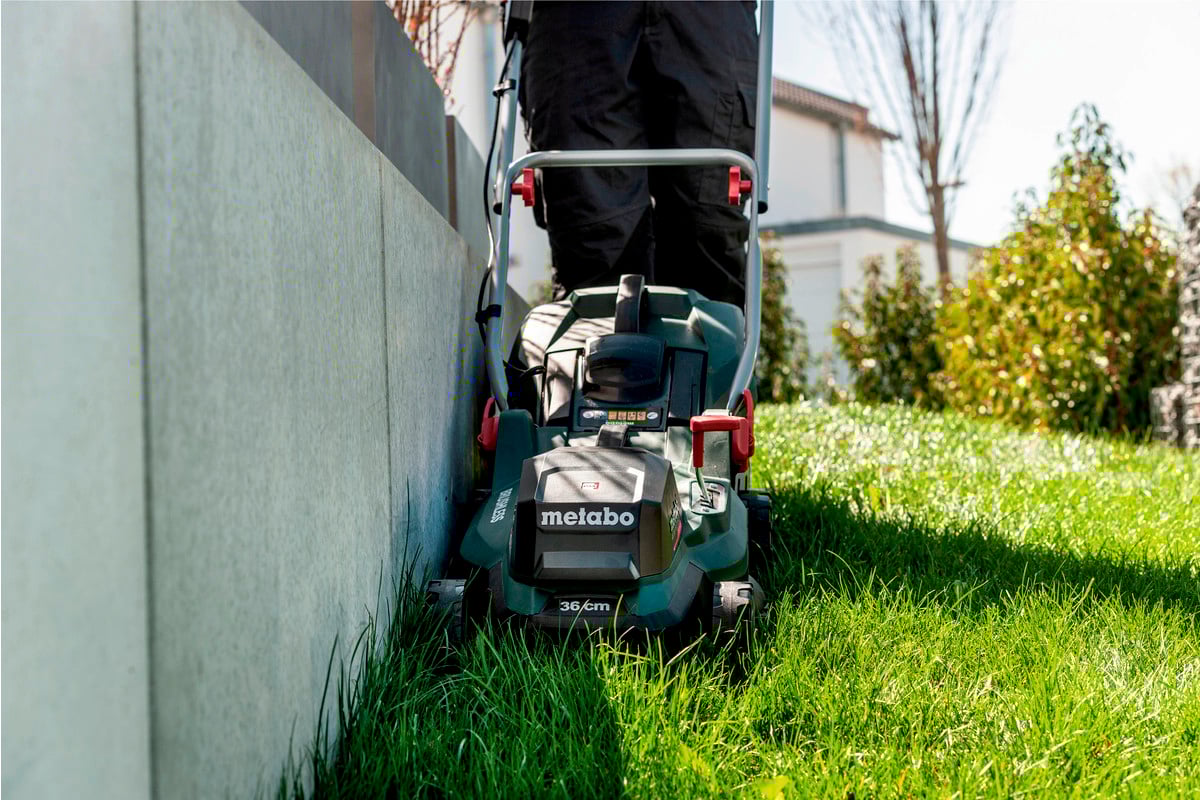 A person is mowing the lawn with a green lawnmower alongside a concrete wall in a sunny garden.
