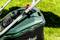 Electric lawn mower on green grass, partially in shade. Handle mechanism and housing section with inscription visible.
