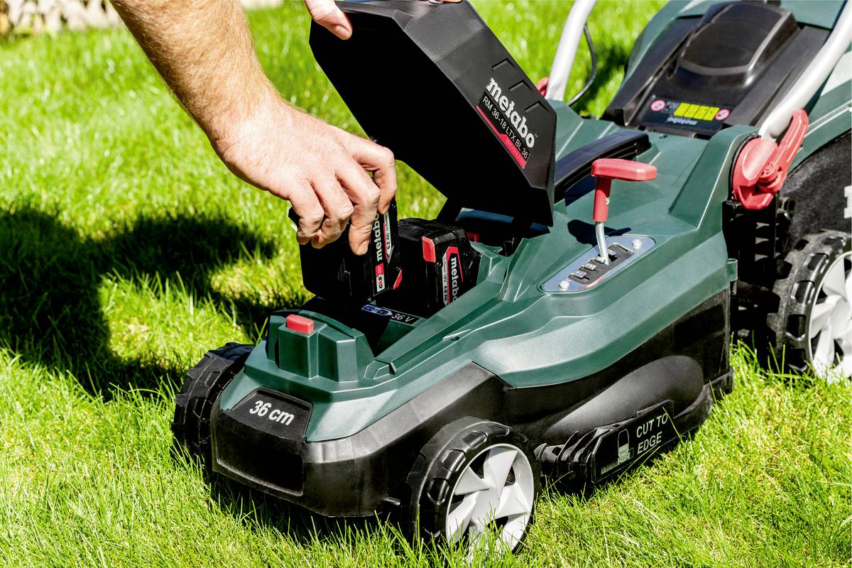 A person is changing the battery on a lawnmower on green grass. The lawnmower displays a '36 cm' label on the front.