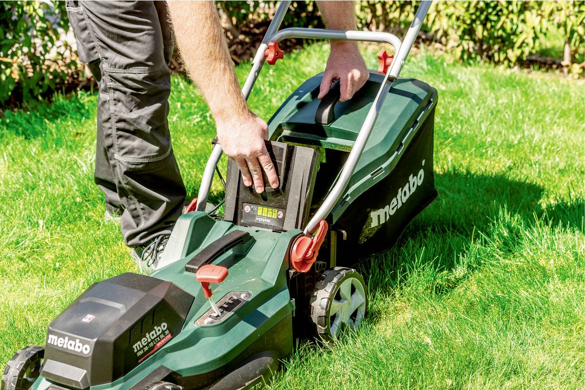 A person is inserting a battery into a green battery-powered lawn mower standing on a grassy area.