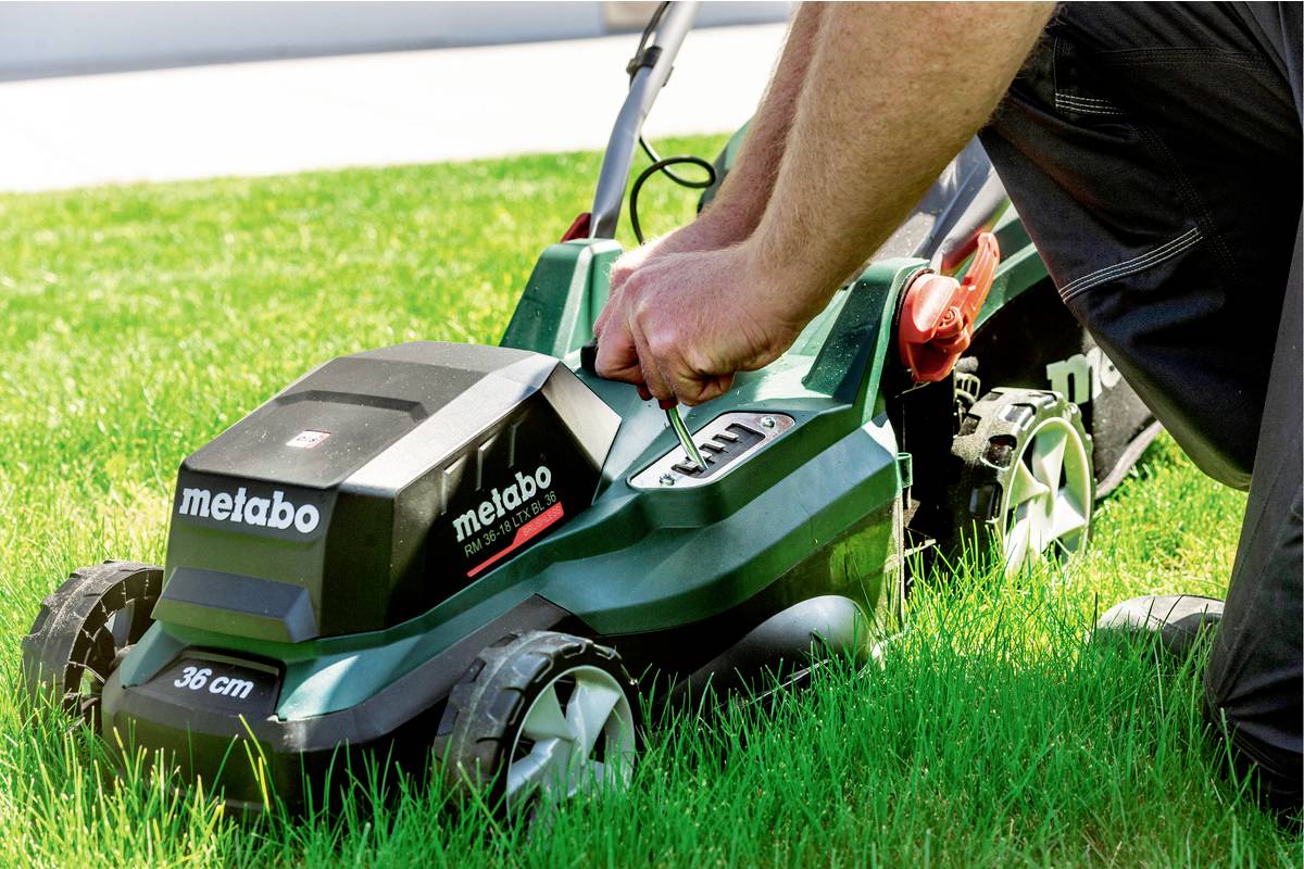 A person is servicing a green lawnmower on a meadow. The lawnmower has the inscription 'metabo'.