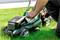 A person is servicing a green lawnmower on a meadow. The lawnmower has the inscription 'metabo'.