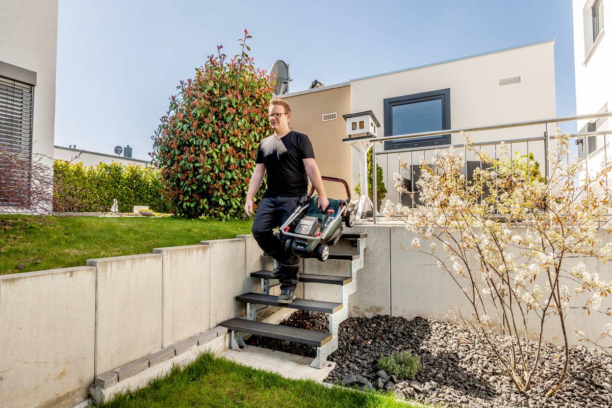 A man is carrying a lawnmower down the stairs in a modern, sunny garden. A house and a bush are visible in the background.