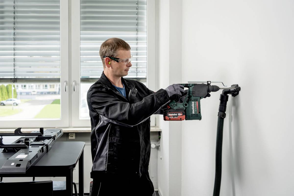 A man wearing safety glasses and ear protection is drilling into a white wall with an electric tool. Windows and a table are visible in the background.