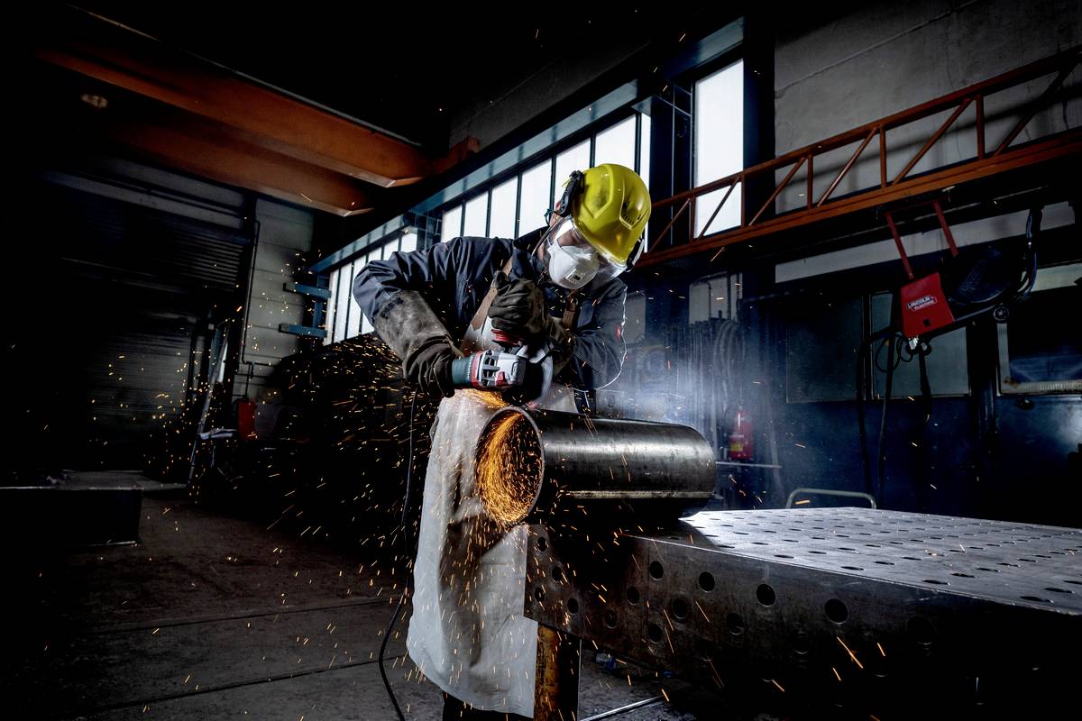 Workers in protective clothing grinding a metal pipe in a factory hall, sparks flying from the grinding tool.