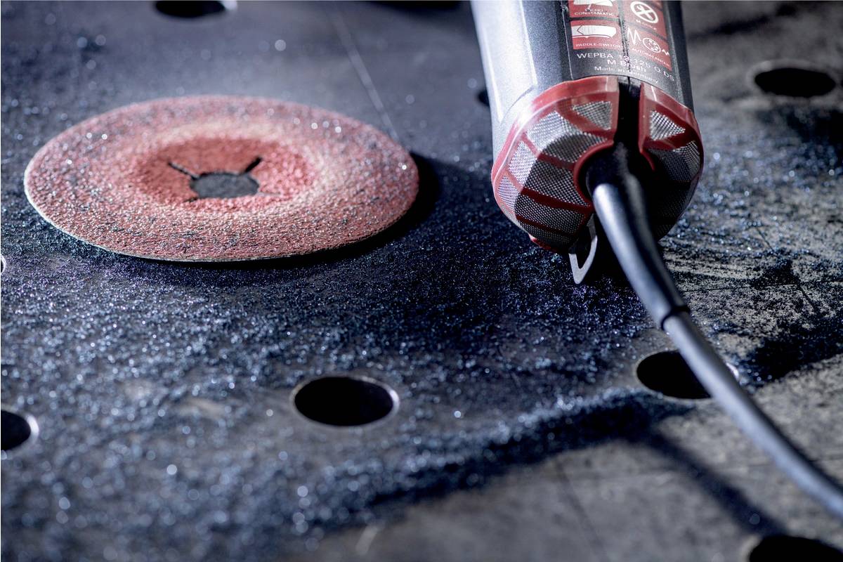 A grinding machine sits beside a red grinding wheel on a metal table with holes, surrounded by grinding dust.