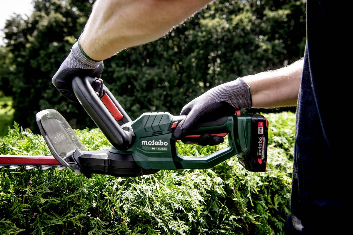 A person is trimming a hedge with an electric hedge trimmer. They are wearing gloves for safety. Dense green vegetation is in the background.