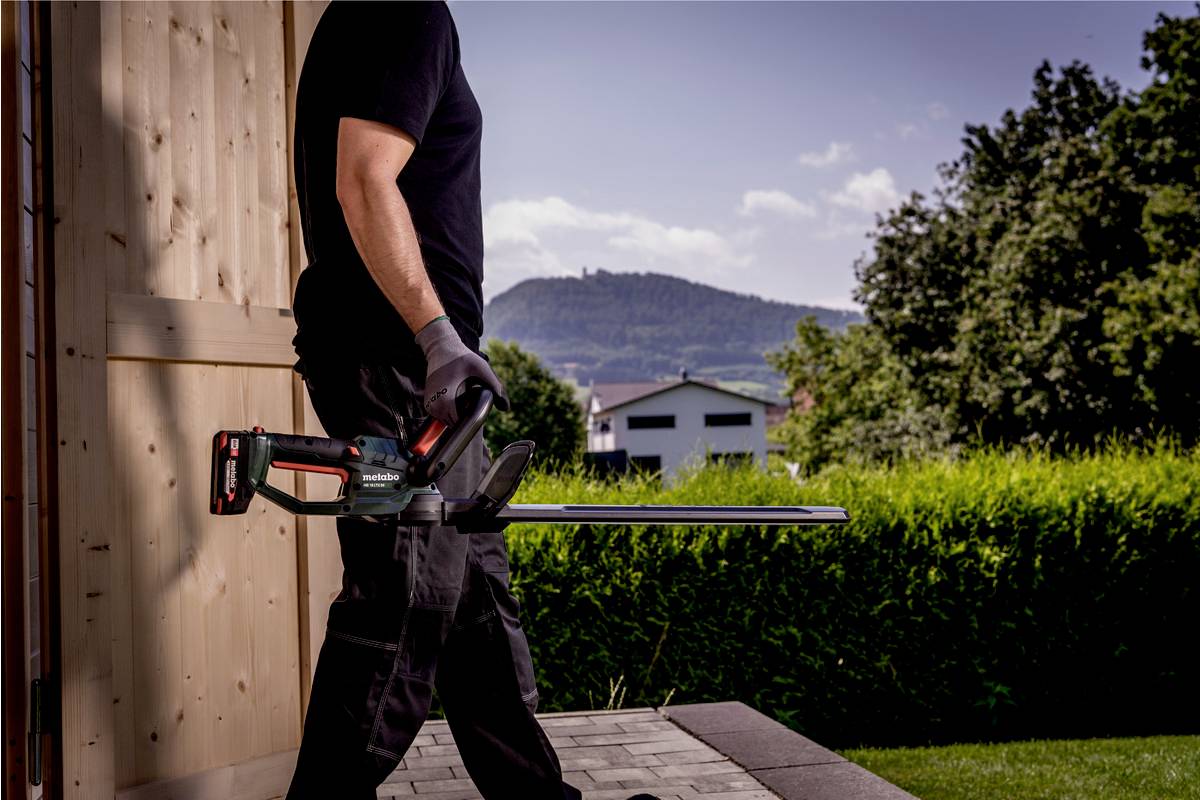A person is carrying a black hedge trimmer outdoors. Green bushes, a building, and a hill are visible in the background.
