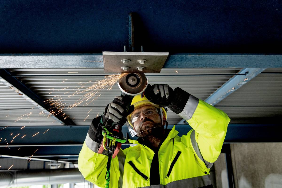 A construction worker in yellow protective clothing is using an angle grinder to process metal beams. Sparks are spraying downwards.