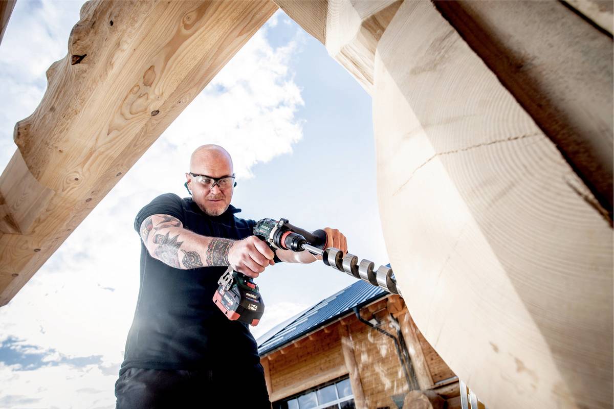 A man with tattoos is using a large cordless drill to bore into wooden beams. Wooden houses can be seen in the background.